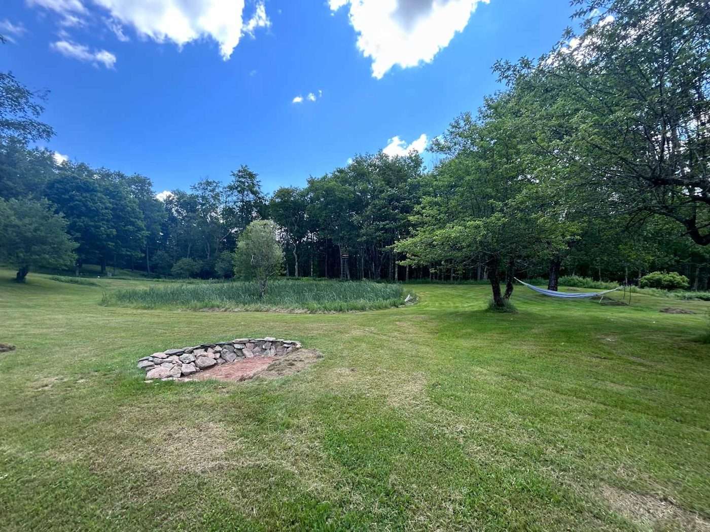 A stone fire ring on the mown field beside the pond at Namaste Catskills, with apple trees and woods behind